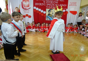 Na tle biało czerwonej dekoracji stoi dziewczynka przebrana za Polskę, obok niej stoi trzech chłopców.